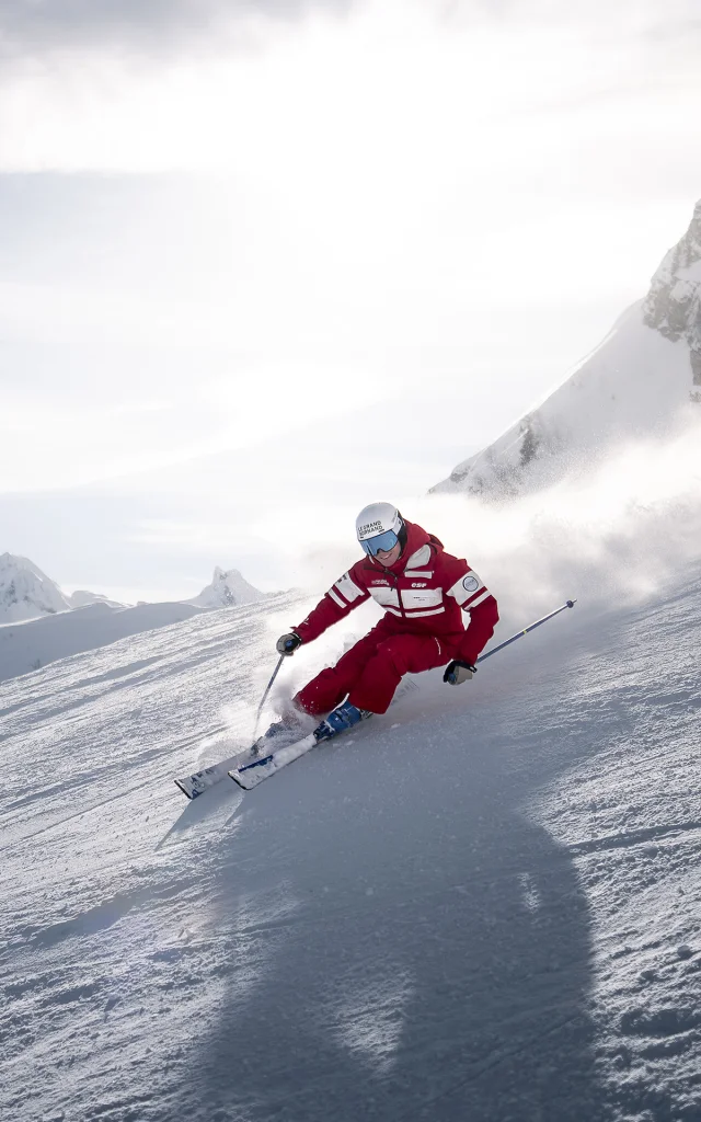 An ESF downhill ski instructor is skiing down a slope with a smile on his face, leaving a cloud of powder behind him. He's wearing a red ski suit in the ESF colours. We can see the Aravis mountain range, with a slightly cloudy blue sky in the background. The atmosphere is both dynamic and gentle.