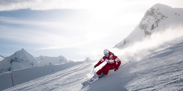 ESF-Skilehrer fährt mit einem Lächeln eine Skipiste hinunter und hinterlässt eine Wolke aus Pulverschnee. Er trägt einen roten Skianzug, der in den Farben der ESF gehalten ist. Wir sehen die Aravis-Kette und einen blauen, leicht bewölkten Himmel im Hintergrund. Die Atmosphäre ist dynamisch und sanft zugleich.