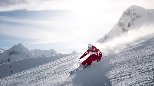 An ESF downhill ski instructor is skiing down a slope with a smile on his face, leaving a cloud of powder behind him. He's wearing a red ski suit in the ESF colours. We can see the Aravis mountain range, with a slightly cloudy blue sky in the background. The atmosphere is both dynamic and gentle.