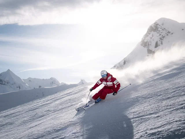 ESF-Skilehrer fährt mit einem Lächeln eine Skipiste hinunter und hinterlässt eine Wolke aus Pulverschnee. Er trägt einen roten Skianzug, der in den Farben der ESF gehalten ist. Wir sehen die Aravis-Kette und einen blauen, leicht bewölkten Himmel im Hintergrund. Die Atmosphäre ist dynamisch und sanft zugleich.