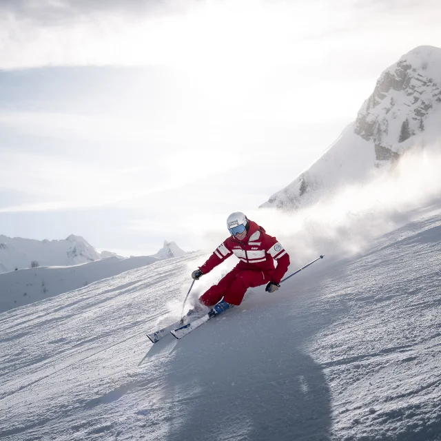 An ESF downhill ski instructor is skiing down a slope with a smile on his face, leaving a cloud of powder behind him. He's wearing a red ski suit in the ESF colours. We can see the Aravis mountain range, with a slightly cloudy blue sky in the background. The atmosphere is both dynamic and gentle.