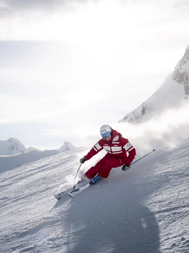 Moniteur de ski alpin de l'ESF dévale une piste de ski avec le sourire laissant derrière lui un nuage de poudreuse. Il est vêtu d'un ensemble de ski rouge, aux couleurs de l'ESF. Nous apercevons la chaîne des Aravis, un ciel bleu un peu nuageux en arrière-plan. L'atmosphère est dynamique et douce à la fois.
