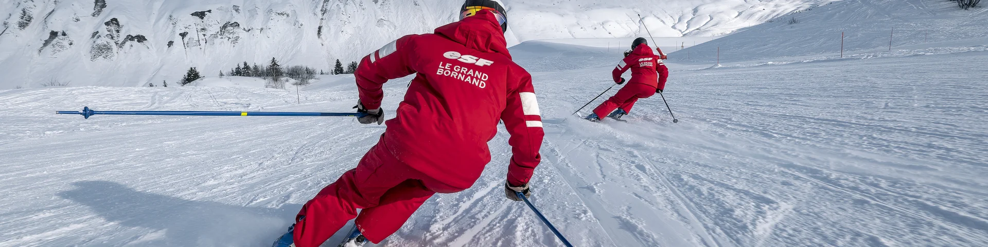 Deux moniteurs de ski alpin de l'ESF descendent une piste de ski plutôt doucement. Ils sont vêtus d'un ensemble de ski rouge, aux couleurs de l'ESF. Nous apercevons la chaîne des Aravis, un ciel bleu un peu nuageux en arrière-plan. L'atmosphère est dynamique et douce à la fois.