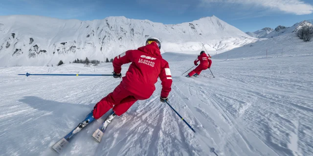 Deux moniteurs de ski alpin de l'ESF descendent une piste de ski plutôt doucement. Ils sont vêtus d'un ensemble de ski rouge, aux couleurs de l'ESF. Nous apercevons la chaîne des Aravis, un ciel bleu un peu nuageux en arrière-plan. L'atmosphère est dynamique et douce à la fois.