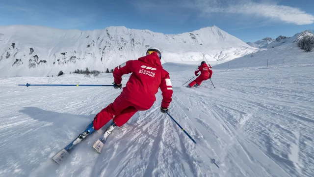 Two ESF downhill ski instructors are skiing down a slope rather slowly. They are dressed in a red ski suit, in the ESF colours. We can see the Aravis mountain range and a slightly cloudy blue sky in the background. The atmosphere is both dynamic and gentle.