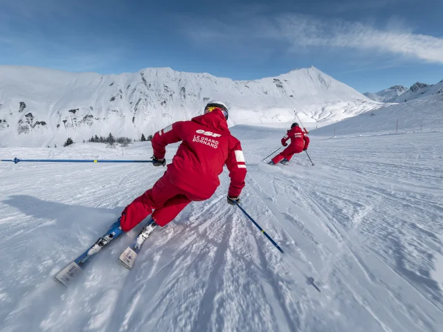 Deux moniteurs de ski alpin de l'ESF descendent une piste de ski plutôt doucement. Ils sont vêtus d'un ensemble de ski rouge, aux couleurs de l'ESF. Nous apercevons la chaîne des Aravis, un ciel bleu un peu nuageux en arrière-plan. L'atmosphère est dynamique et douce à la fois.