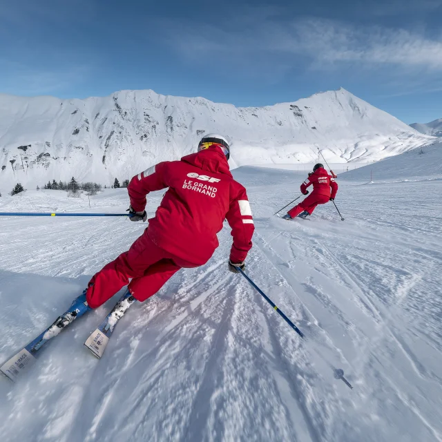 Deux moniteurs de ski alpin de l'ESF descendent une piste de ski plutôt doucement. Ils sont vêtus d'un ensemble de ski rouge, aux couleurs de l'ESF. Nous apercevons la chaîne des Aravis, un ciel bleu un peu nuageux en arrière-plan. L'atmosphère est dynamique et douce à la fois.