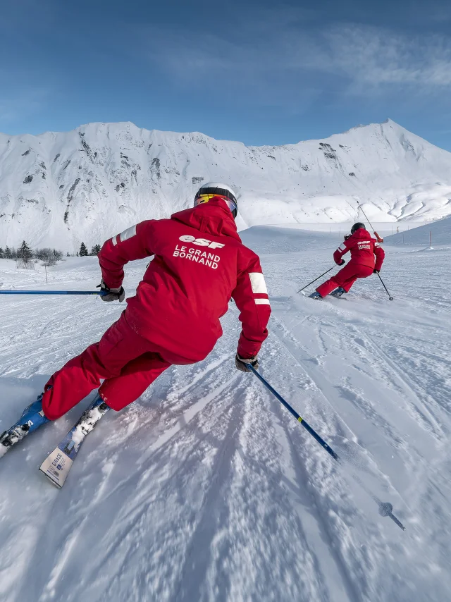 Deux moniteurs de ski alpin de l'ESF descendent une piste de ski plutôt doucement. Ils sont vêtus d'un ensemble de ski rouge, aux couleurs de l'ESF. Nous apercevons la chaîne des Aravis, un ciel bleu un peu nuageux en arrière-plan. L'atmosphère est dynamique et douce à la fois.