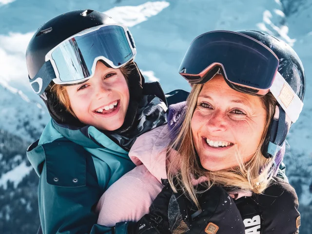 Une mère de famille portant son enfant sur son dos. Ils portent tous les deux des vêtements et des équipements de ski et sourient à la caméra. Nous pouvons voir la chaîne des Aravis enneigée avec la pointe Percée en arrière-plan. L'image évoque un sentiment de joie, d'aventure et de lien familial, typique d'une journée de ski en famille. La composition est chaleureuse et met l'accent sur le bonheur partagé.