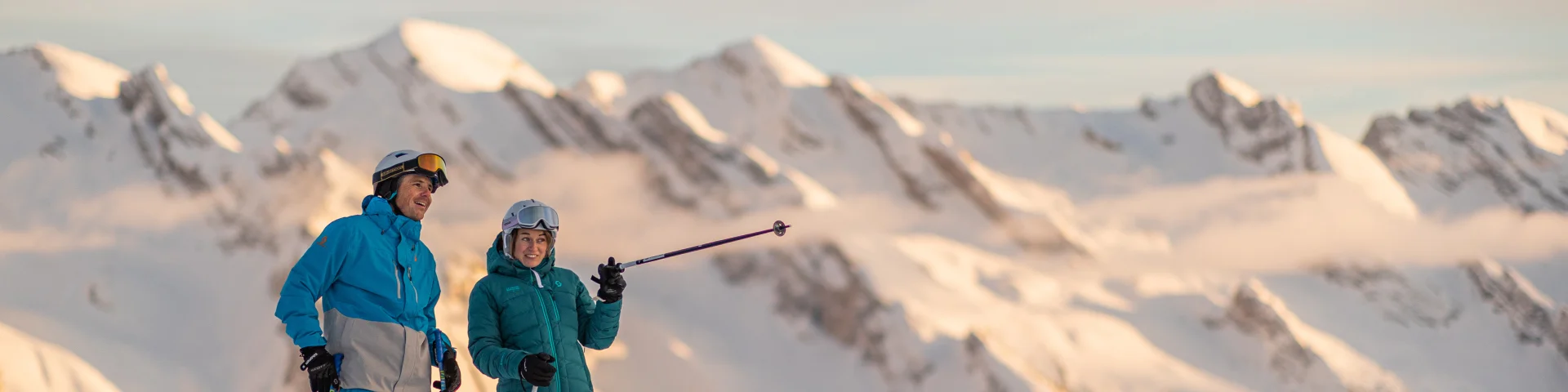 Deux skieurs alpins se sont arrêtés quelques minutes en haut des pistes pour contempler le coucher de soleil et la vue imprenable sur la chaîne des Aravis. L'un est vêtu d'un ensemble de ski bleu et gris et l'autre d'un ensemble de ski bleu turquoise. Une belle complicité se fait ressentir, tout deux souriants. L'un des skieurs montre en levant le bâton, une montagne certainement, à son ami. L'atmosphère est détendue et les lumières assez réconfortantes.