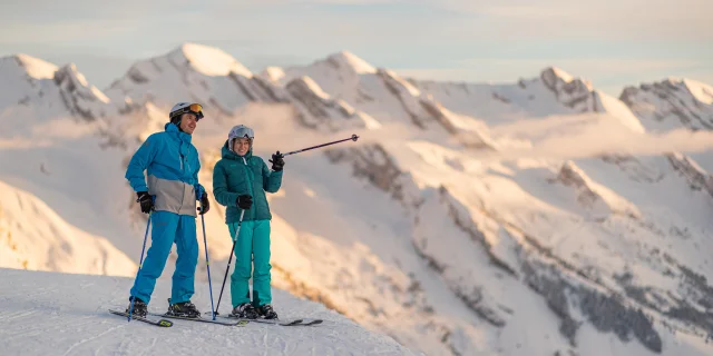 Deux skieurs alpins se sont arrêtés quelques minutes en haut des pistes pour contempler le coucher de soleil et la vue imprenable sur la chaîne des Aravis. L'un est vêtu d'un ensemble de ski bleu et gris et l'autre d'un ensemble de ski bleu turquoise. Une belle complicité se fait ressentir, tout deux souriants. L'un des skieurs montre en levant le bâton, une montagne certainement, à son ami. L'atmosphère est détendue et les lumières assez réconfortantes.