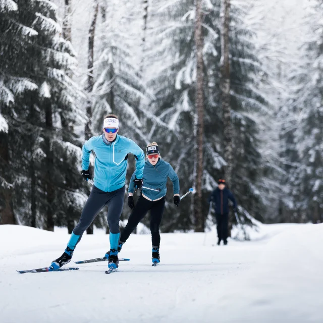 A couple of Nordic skiers are skiing with intensity on the Grand-Bornand Nordic ski area between snow-covered fir trees. The snow is brilliant. The skiers are in the foreground, moving straight ahead. We can see someone walking in the background. The skiers are wearing blue, black and grey Nordic skis with the latest equipment.