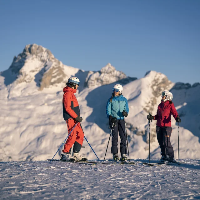 Trois personnes sur des skis, ont un fou-rire sur un sommet enneigé. Cette scène hivernale évoque l'esprit d'aventure et de camaraderie. La chaîne des Aravis et la Pointe Percée en arrière-plan ajoutent une touche de grandeur, tandis que la neige scintillante sous le soleil crée une atmosphère magique.
