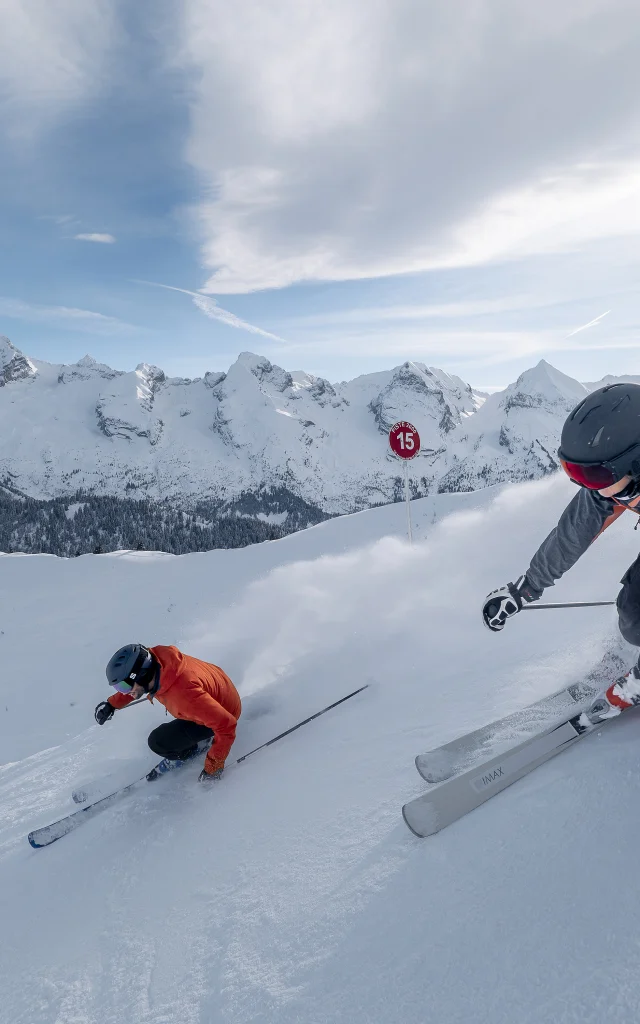 Two skiers hurtle down a snow-covered slope, leaving a cloud of powder behind them. One is wearing a grey and pink jacket with a black helmet, while the other is wearing a red jacket. The Aravis mountain range is in the background, under a partly cloudy blue sky. The scene captures the intensity and adrenalin of skiing.