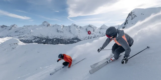 Deux skieurs dévalent une pente enneigée à grande vitesse, laissant derrière eux un nuage de poudreuse. L'un porte une veste grise et rose avec un casque noir, tandis que l'autre est vêtu d'une veste rouge. La chaîne des Aravis est présente en arrière-plan, sous un ciel bleu partiellement nuageux. La scène capture l'intensité et l'adrénaline du ski.