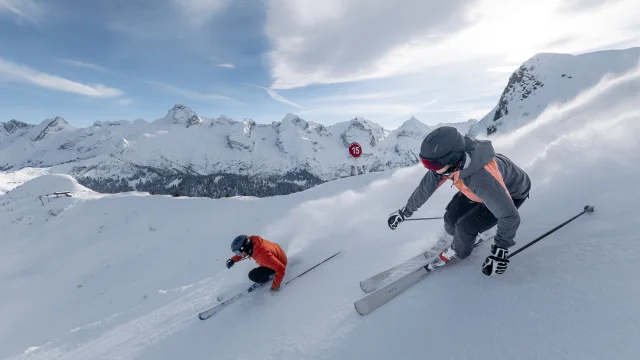 Deux skieurs dévalent une pente enneigée à grande vitesse, laissant derrière eux un nuage de poudreuse. L'un porte une veste grise et rose avec un casque noir, tandis que l'autre est vêtu d'une veste rouge. La chaîne des Aravis est présente en arrière-plan, sous un ciel bleu partiellement nuageux. La scène capture l'intensité et l'adrénaline du ski.