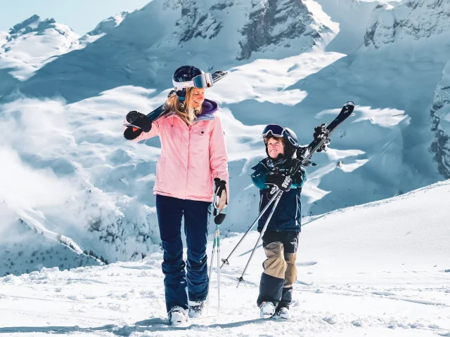Eine Mutter und ihr Sohn, beide im Skianzug (die Mutter in rosa und schwarz mit Helm und Skibrille auf dem Kopf und ihr Sohn in schwarz und braun mit Helm und Skibrille auf dem Kopf), tragen ihre Skier auf ihren Schultern, der kleine Junge hat etwas Mühe, sie zu tragen. Beide schauen sich mit einer sehr schönen Komplizenschaft an, während sie durch den Schnee stapfen. Wir können die Pointe Percée im Hintergrund sehen, mit einem sehr schönen klaren blauen Himmel.