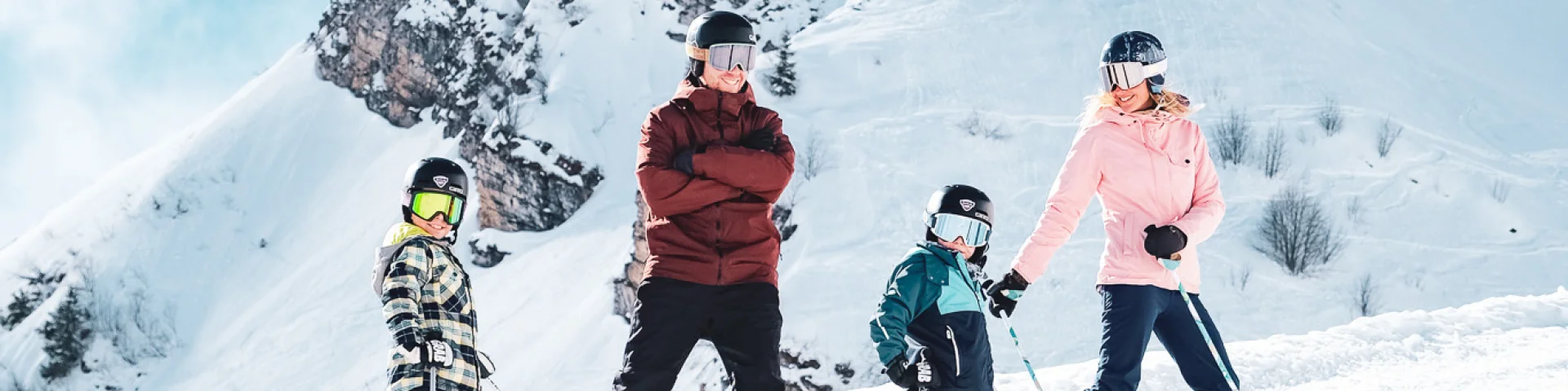 Une famille composée d'un père vêtu d'habits de ski rouge et noire et d'un snowboard, d'une maman avec des habits de ski rose et noire et deux petits garçons avec des habits de ski multicolore, posent en ski devant la Pointe Percée avec un grand sourire. Ils sont prêts à descendre la piste. Une belle atmosphère ce dégage de cette image avec une neige blanche et un ciel bleu un peu nuageux.