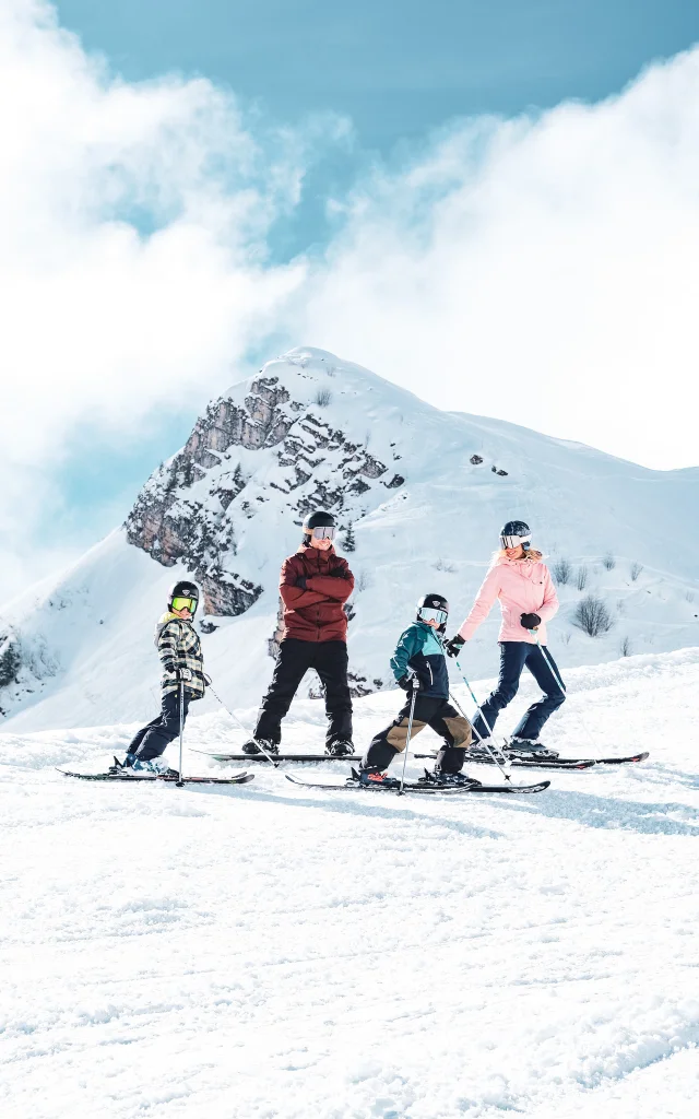 A family consisting of a father in red and black ski clothes and a snowboard, a mother in pink and black ski clothes and two little boys in multicoloured ski clothes, pose on skis in front of the Pointe Percée with big smiles on their faces. They're ready to hit the slopes. The image has a lovely atmosphere, with white snow and a slightly cloudy blue sky.