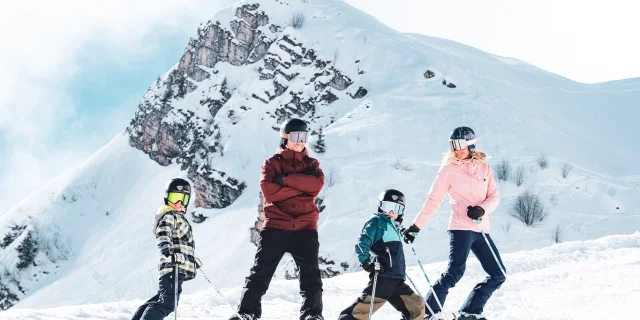 Une famille composée d'un père vêtu d'habits de ski rouge et noire et d'un snowboard, d'une maman avec des habits de ski rose et noire et deux petits garçons avec des habits de ski multicolore, posent en ski devant la Pointe Percée avec un grand sourire. Ils sont prêts à descendre la piste. Une belle atmosphère ce dégage de cette image avec une neige blanche et un ciel bleu un peu nuageux.