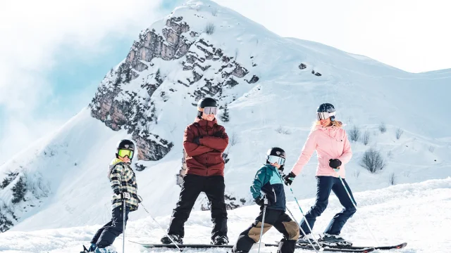 Eine Familie, bestehend aus einem Vater in rot-schwarzer Skikleidung und einem Snowboard, einer Mutter in rot-schwarzer Skikleidung und zwei kleinen Jungen in bunten Skikleidern, posiert mit einem breiten Lächeln auf Skiern vor der Pointe Percée. Sie sind bereit, die Piste hinunterzufahren. Das Bild vermittelt eine schöne Atmosphäre mit weißem Schnee und einem blauen, leicht bewölkten Himmel.