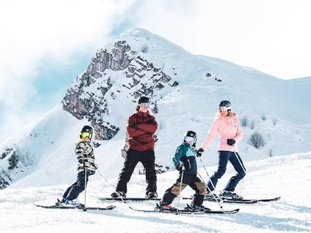 A family consisting of a father in red and black ski clothes and a snowboard, a mother in pink and black ski clothes and two little boys in multicoloured ski clothes, pose on skis in front of the Pointe Percée with big smiles on their faces. They're ready to hit the slopes. The image has a lovely atmosphere, with white snow and a slightly cloudy blue sky.