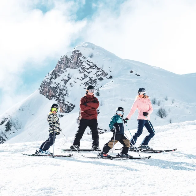 A family consisting of a father in red and black ski clothes and a snowboard, a mother in pink and black ski clothes and two little boys in multicoloured ski clothes, pose on skis in front of the Pointe Percée with big smiles on their faces. They're ready to hit the slopes. The image has a lovely atmosphere, with white snow and a slightly cloudy blue sky.