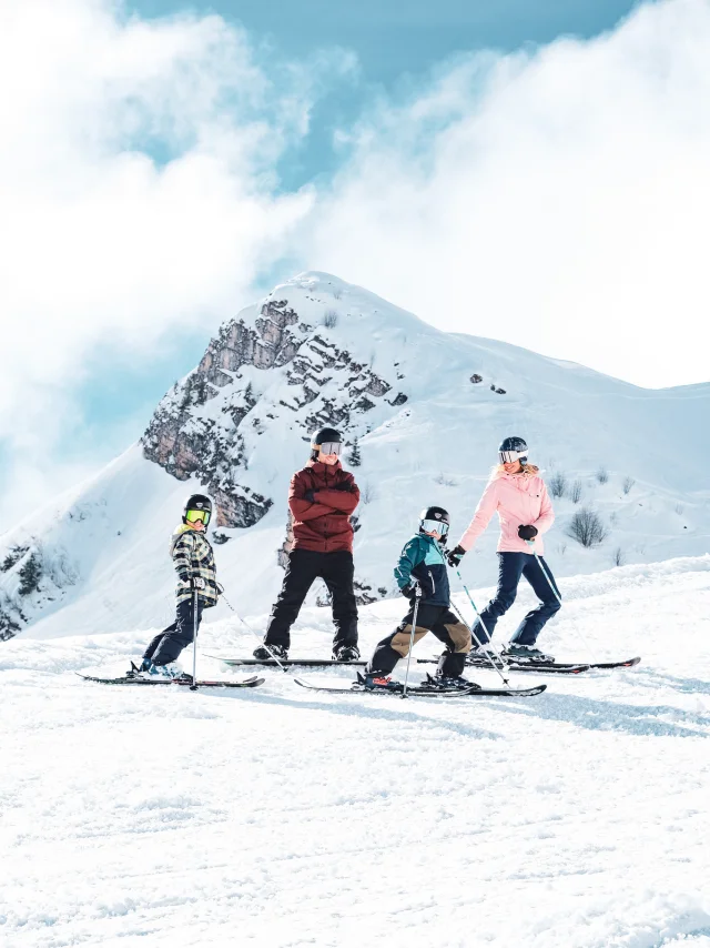 A family consisting of a father in red and black ski clothes and a snowboard, a mother in pink and black ski clothes and two little boys in multicoloured ski clothes, pose on skis in front of the Pointe Percée with big smiles on their faces. They're ready to hit the slopes. The image has a lovely atmosphere, with white snow and a slightly cloudy blue sky.