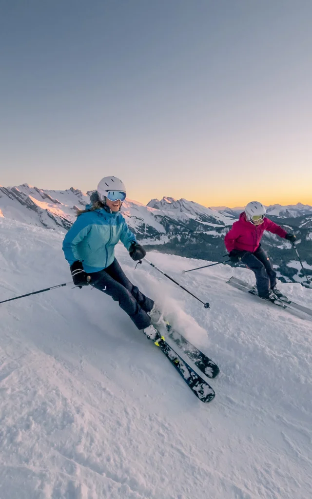 Deux skieuses dévalant une pente enneigée au coucher du soleil. Cette scène magique capture la beauté des paysages d'hiver, où les flocons de neige scintillent sous les rayons dorés du soleil couchant. Les skieuses, vêtues d'un ensemble de ski bleu et rose, semblent s'amuser tout en maîtrisant leur descente, créant une atmosphère de joie et d'aventure. Nous pouvons voir la chaîne des Aravis en arrière-plan. Le contraste entre le blanc éclatant de la neige et les teintes chaudes du ciel ajoute une dimension visuelle saisissante, invitant à la rêverie et à l'évasion.