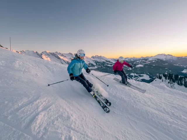 Deux skieuses dévalant une pente enneigée au coucher du soleil. Cette scène magique capture la beauté des paysages d'hiver, où les flocons de neige scintillent sous les rayons dorés du soleil couchant. Les skieuses, vêtues d'un ensemble de ski bleu et rose, semblent s'amuser tout en maîtrisant leur descente, créant une atmosphère de joie et d'aventure. Nous pouvons voir la chaîne des Aravis en arrière-plan. Le contraste entre le blanc éclatant de la neige et les teintes chaudes du ciel ajoute une dimension visuelle saisissante, invitant à la rêverie et à l'évasion.