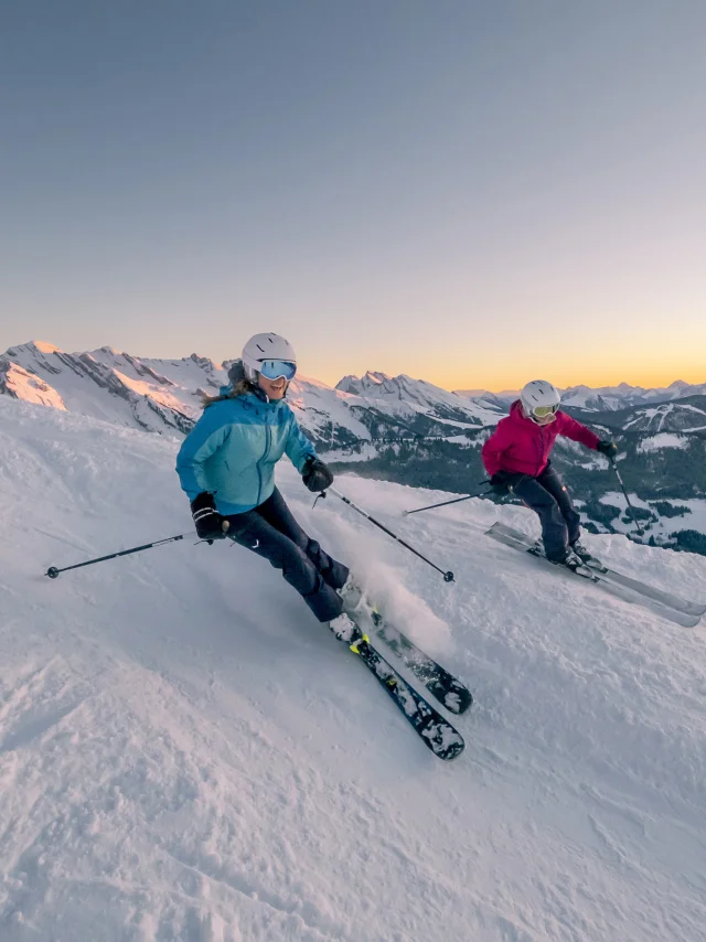 Deux skieuses dévalant une pente enneigée au coucher du soleil. Cette scène magique capture la beauté des paysages d'hiver, où les flocons de neige scintillent sous les rayons dorés du soleil couchant. Les skieuses, vêtues d'un ensemble de ski bleu et rose, semblent s'amuser tout en maîtrisant leur descente, créant une atmosphère de joie et d'aventure. Nous pouvons voir la chaîne des Aravis en arrière-plan. Le contraste entre le blanc éclatant de la neige et les teintes chaudes du ciel ajoute une dimension visuelle saisissante, invitant à la rêverie et à l'évasion.