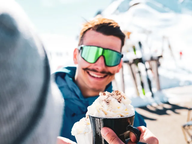 Un jeune homme avec une moustache en habits de ski bleu canard et avec des lunettes de soleil à verre vert en arrière-plan sourit à son amie en trinquant tous deux avec leur chocolat chaud avec chantilly. Cette scène en haut des pistes montre un vrai moment de complicité et de gourmandise.