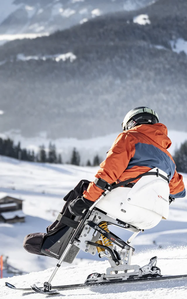 Eine Person mit eingeschränkter Mobilität betreibt Handskifahren in einer verschneiten Umgebung. Der Skifahrer trägt einen schwarzen, orangefarbenen und grauen Skianzug und einen schwarzen Helm. Im Hintergrund können wir das Bargy-Massiv erkennen. Die Atmosphäre ist sehr sanft und heiter.
