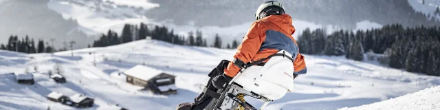 Une personne à mobilité réduite pratique le handiski dans un décor enneigé. Le skieur est vêtu d'un ensemble de ski noir, orange et gris avec un casque noir. Nous pouvons apercevoir le massif du Bargy en arrière-plan. L'atmosphère est très douce et serène.