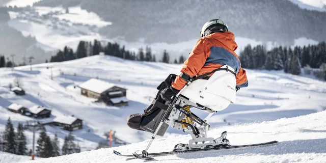 Une personne à mobilité réduite pratique le handiski dans un décor enneigé. Le skieur est vêtu d'un ensemble de ski noir, orange et gris avec un casque noir. Nous pouvons apercevoir le massif du Bargy en arrière-plan. L'atmosphère est très douce et serène.
