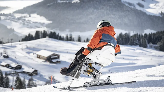 Eine Person mit eingeschränkter Mobilität betreibt Handskifahren in einer verschneiten Umgebung. Der Skifahrer trägt einen schwarzen, orangefarbenen und grauen Skianzug und einen schwarzen Helm. Im Hintergrund können wir das Bargy-Massiv erkennen. Die Atmosphäre ist sehr sanft und heiter.