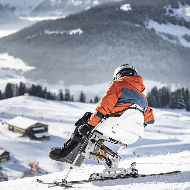 A person with reduced mobility goes handiskiing in a snowy setting. The skier is wearing a black, orange and grey ski suit with a black helmet. We can see the Bargy massif in the background. The atmosphere is soft and serene.