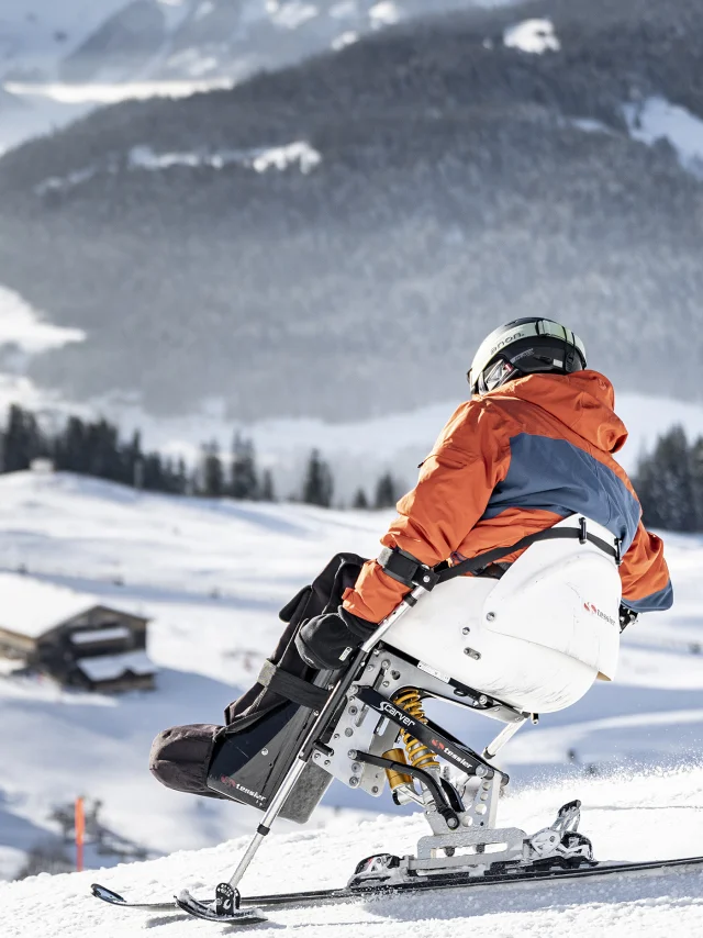 Une personne à mobilité réduite pratique le handiski dans un décor enneigé. Le skieur est vêtu d'un ensemble de ski noir, orange et gris avec un casque noir. Nous pouvons apercevoir le massif du Bargy en arrière-plan. L'atmosphère est très douce et serène.