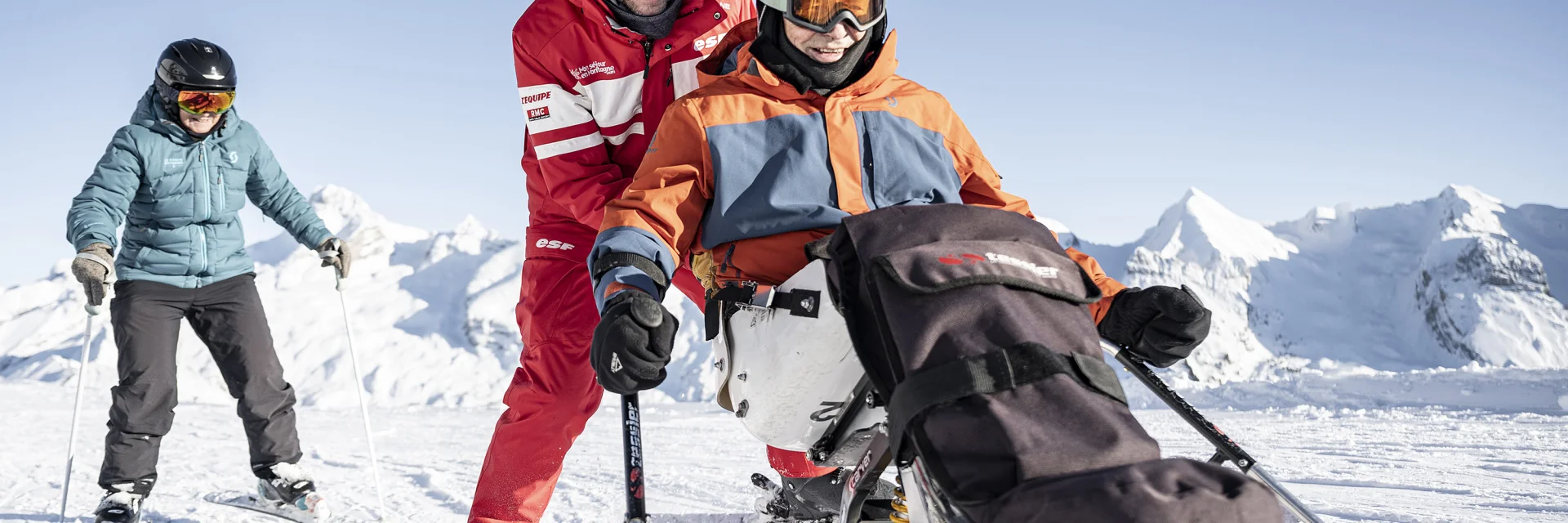 A person with reduced mobility goes handiskiing in a snowy setting. His ESF guide in red clothes pushes and belays behind him, both laughing and seeming to enjoy the moment. Behind them, the PRM skier's wife follows them. The PRM skier is wearing a black, orange and grey ski suit with a black helmet. We can see the Aravis mountain range in the background. The atmosphere is very gentle, serene and jovial.