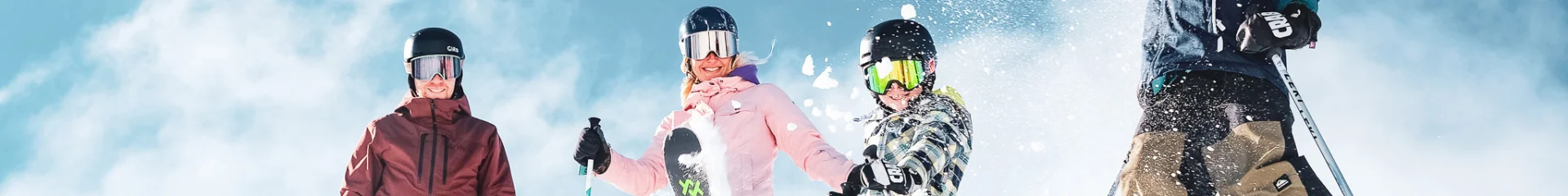 Une famille de quatre personnes en tenue de ski colorées ainsi que des équipements de ski comprenant des gants, des casques et des lunettes de protection, dans un environnement enneigé sous un ciel bleu clair. Les personnes sont placées en haut d'une pente enneigée. Un enfant au centre projette de la neige en l'air avec son ski, créant une dynamique joyeuse et animée.