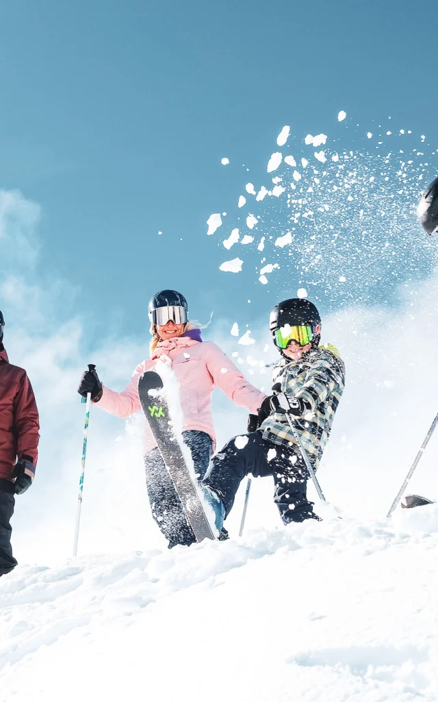 Eine vierköpfige Familie in bunten Skianzügen sowie einer Skiausrüstung, zu der Handschuhe, Helme und Schutzbrillen gehören, in einer verschneiten Umgebung unter einem hellblauen Himmel. Die Personen werden oben auf einem schneebedeckten Hang platziert. Ein Kind in der Mitte schleudert mit seinem Ski Schnee in die Luft, wodurch eine fröhliche und lebhafte Dynamik entsteht.