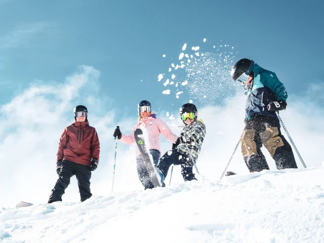 Une famille de quatre personnes en tenue de ski colorées ainsi que des équipements de ski comprenant des gants, des casques et des lunettes de protection, dans un environnement enneigé sous un ciel bleu clair. Les personnes sont placées en haut d'une pente enneigée. Un enfant au centre projette de la neige en l'air avec son ski, créant une dynamique joyeuse et animée.