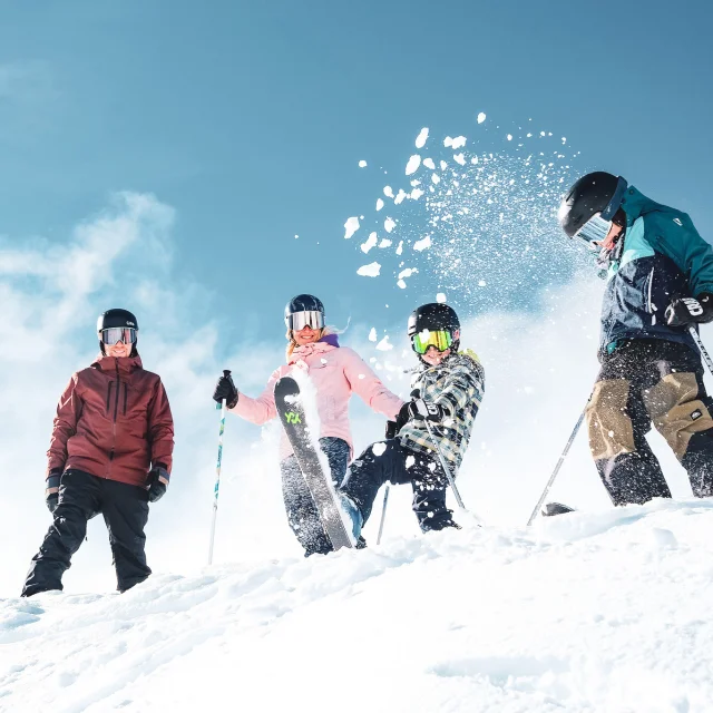 Une famille de quatre personnes en tenue de ski colorées ainsi que des équipements de ski comprenant des gants, des casques et des lunettes de protection, dans un environnement enneigé sous un ciel bleu clair. Les personnes sont placées en haut d'une pente enneigée. Un enfant au centre projette de la neige en l'air avec son ski, créant une dynamique joyeuse et animée.