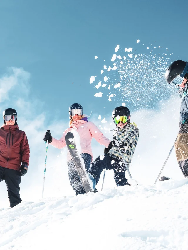 Une famille de quatre personnes en tenue de ski colorées ainsi que des équipements de ski comprenant des gants, des casques et des lunettes de protection, dans un environnement enneigé sous un ciel bleu clair. Les personnes sont placées en haut d'une pente enneigée. Un enfant au centre projette de la neige en l'air avec son ski, créant une dynamique joyeuse et animée.