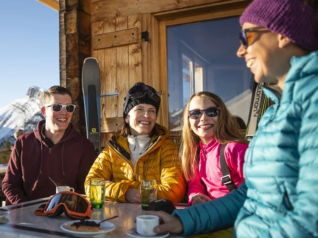 Groupe d'amis avec une enfant se retrouve autour d'une table en plein air, profitant d'un moment convivial après une journée de ski. Les sourires radieux et les rires partagés témoignent de l'esprit chaleureux qui règne dans cette ambiance montagnarde. La chaîne des Aravis en arrière-plan ajoute une touche de sérénité, tandis que les chaises en bois rustiques et la table en plein air évoquent une atmosphère accueillante et décontractée. Que ce soit pour déguster un chocolat chaud, une tarte à la myrtille, un café ou partager des anecdotes de ski, cette scène illustre parfaitement le plaisir de se rassembler en pleine nature.
