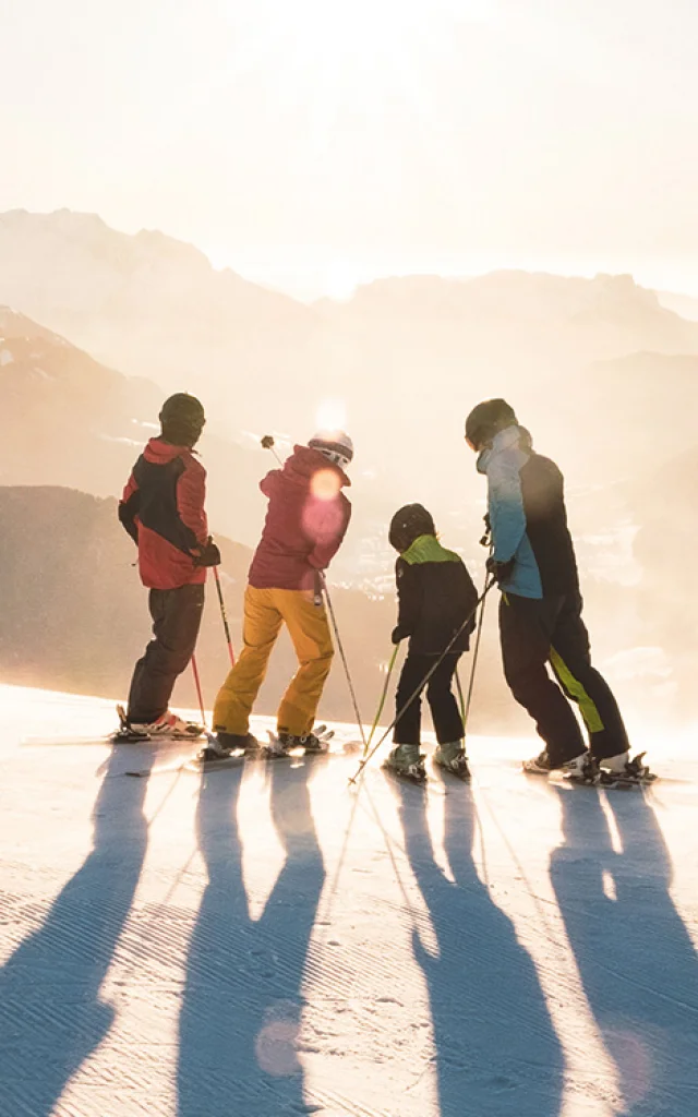 A family with two children standing on skis at the top of a slope watching the sun set over the Aravis mountain range. One has a blue, black and green ski outfit, another red and yellow, another green and black and the last red and black. One of the skiers in the middle is pointing with his left stick to the top of a mountain. The atmosphere is very lunar with a white/golden light. It's a beautiful end to a day's skiing.