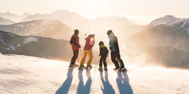 A family with two children standing on skis at the top of a slope watching the sun set over the Aravis mountain range. One has a blue, black and green ski outfit, another red and yellow, another green and black and the last red and black. One of the skiers in the middle is pointing with his left stick to the top of a mountain. The atmosphere is very lunar with a white/golden light. It's a beautiful end to a day's skiing.