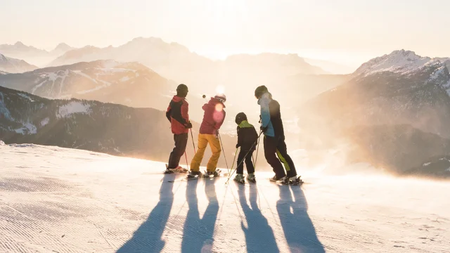 Eine Familie mit zwei Kindern steht auf Skiern am oberen Ende einer Piste und betrachtet den Sonnenuntergang mit Blick auf die Aravis-Kette. Einer hat einen blau-schwarz-grünen Skisatz, ein anderer einen rot-gelben, ein weiterer einen grün-schwarzen und der letzte einen rot-schwarzen. Einer der Skifahrer in der Mitte zeigt mit seinem linken Stock auf den Gipfel eines Berges. Die Atmosphäre ist sehr mondähnlich mit weißem/goldenem Licht. Es ist ein schöner Abschluss eines Skitages.