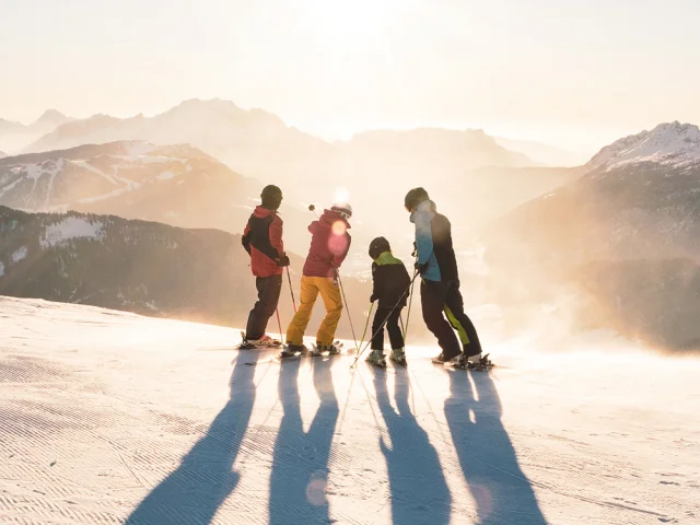 Une famille composée de deux enfants se tenant sur des skis en haut d'une piste contemple le coucher de soleil avec vue sur la chaîne des Aravis. L'un a un ensemble de ski bleu, noir et vert, un autre rouge et jaune, un autre vert et noir et le dernier rouge et noir. L'un des skieurs au milieu pointe avec son bâton gauche le sommet d'une montagne. L'atmosphère est très lunaire avec une lumière blanche/dorée. C'est une belle fin de journée de ski.