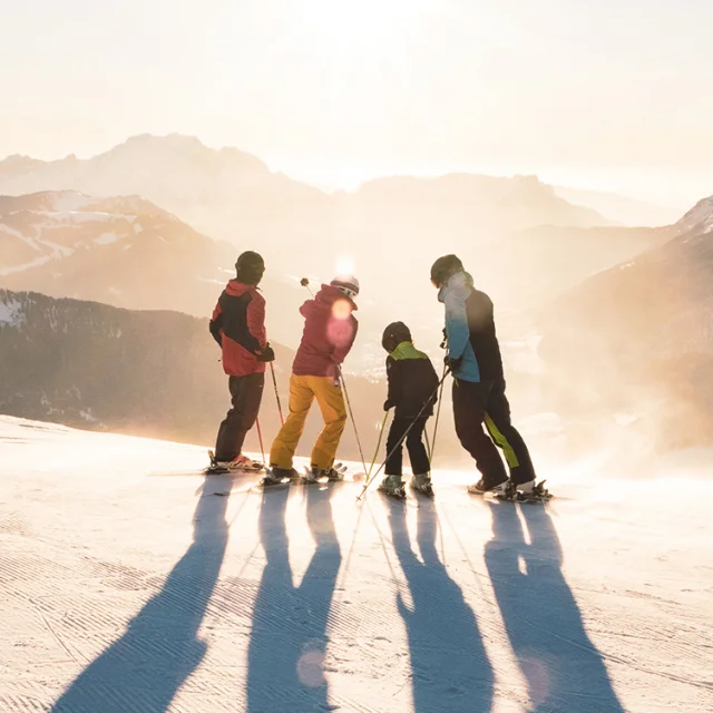 A family with two children standing on skis at the top of a slope watching the sun set over the Aravis mountain range. One has a blue, black and green ski outfit, another red and yellow, another green and black and the last red and black. One of the skiers in the middle is pointing with his left stick to the top of a mountain. The atmosphere is very lunar with a white/golden light. It's a beautiful end to a day's skiing.